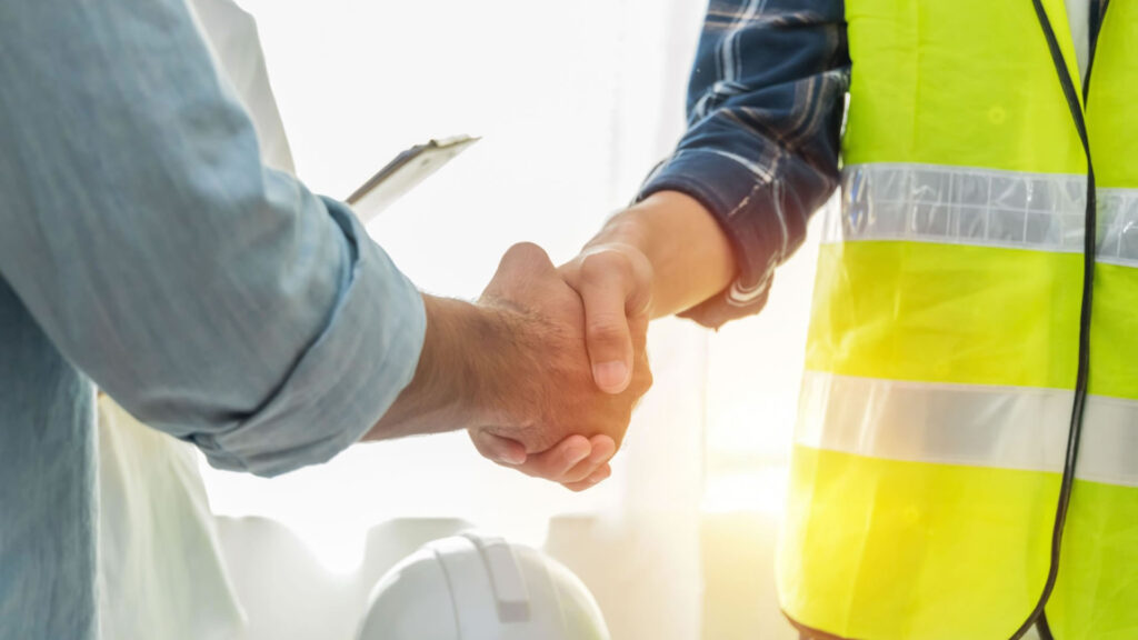 Two construction workers in hard hats shaking hands on a job site, symbolizing teamwork and collaboration.