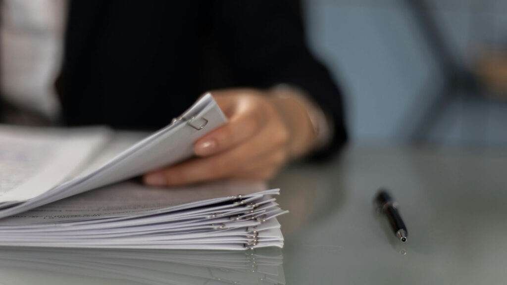 A person in a suit stands holding a stack of papers, looking focused and ready to present information.